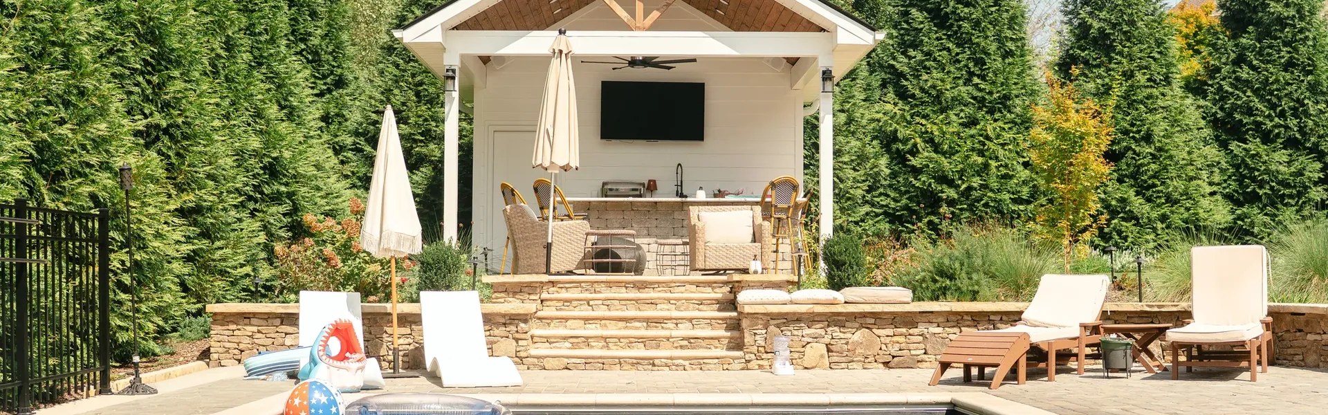 Covered outdoor kitchen pavilion with a stone bar island, bar stools and a mounted television, above dry-stacked stone steps and a dark-bottom pool in Madison, Alabama