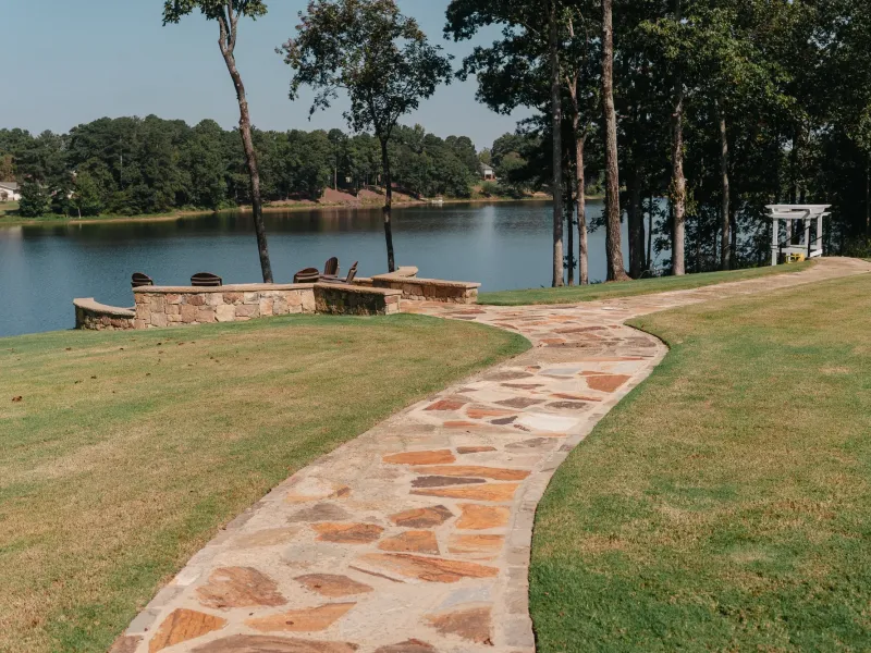 Mortared flagstone walkway curving across a lawn to a lakeside stone seat wall and fire pit with Adirondack chairs, on Lake Catoma in Cullman, Alabama