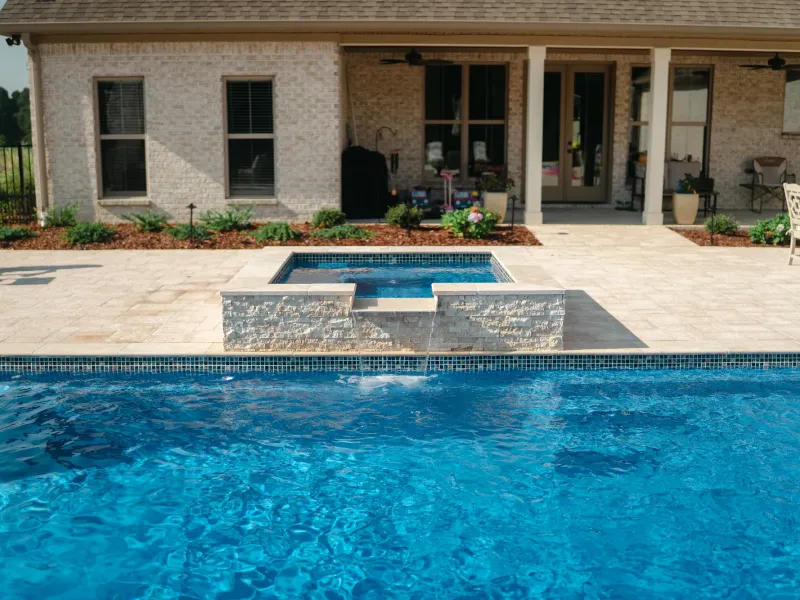 Rectangular pool with a square spillover spa and stacked stone water feature on a travertine deck in Guntersville, Alabama