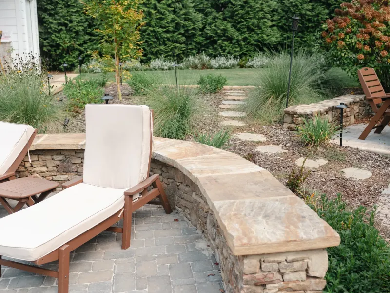 Curved dry-stacked stone seat wall with a thick sandstone cap edging a cobblestone paver patio, with teak loungers and ornamental grasses in Madison, Alabama