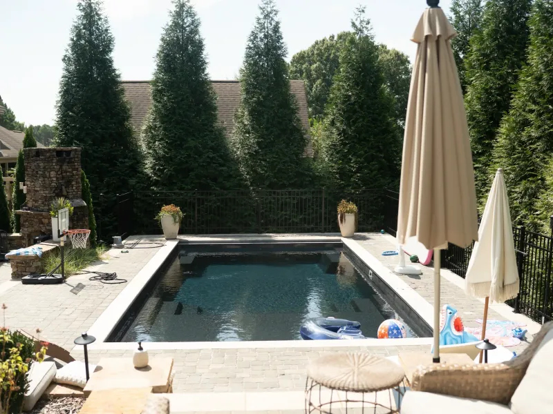 Dark-bottom rectangular pool with a paver deck, patio umbrellas and a stacked stone outdoor fireplace beyond, screened by arborvitae in Madison, Alabama