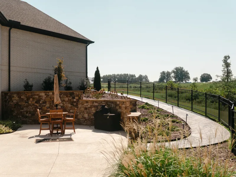 Stamped concrete walkway curving past a mortared stone retaining wall and ornamental grasses to a concrete patio with bar seating and a grill in Cullman, Alabama