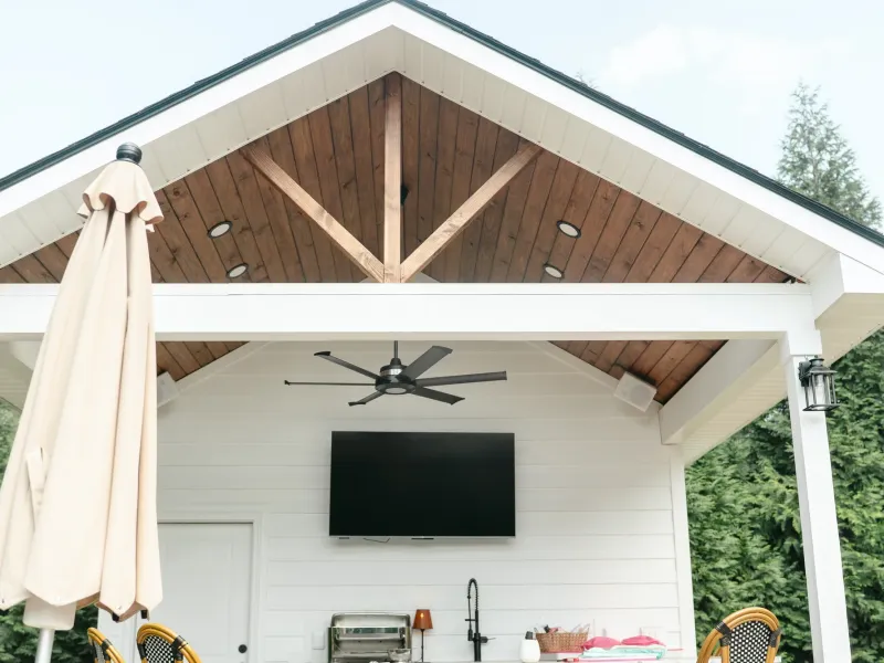 Gabled outdoor kitchen pavilion with an exposed cedar ceiling, ceiling fan, mounted television and a stone bar island in Madison, Alabama