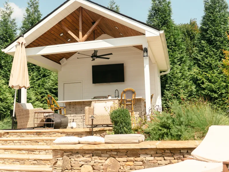 Outdoor kitchen under a gabled pavilion with a stacked block island, stone counter, bar stools and a sink tap, above dry-stacked stone steps in Madison, Alabama